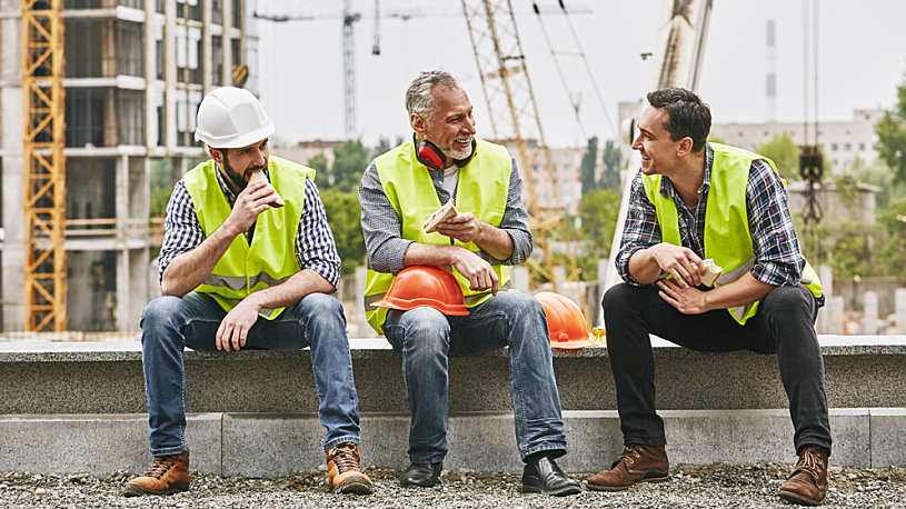 Construction Industry Trends Three construction coworkers sit on a low stone wall and talk while eating during a break