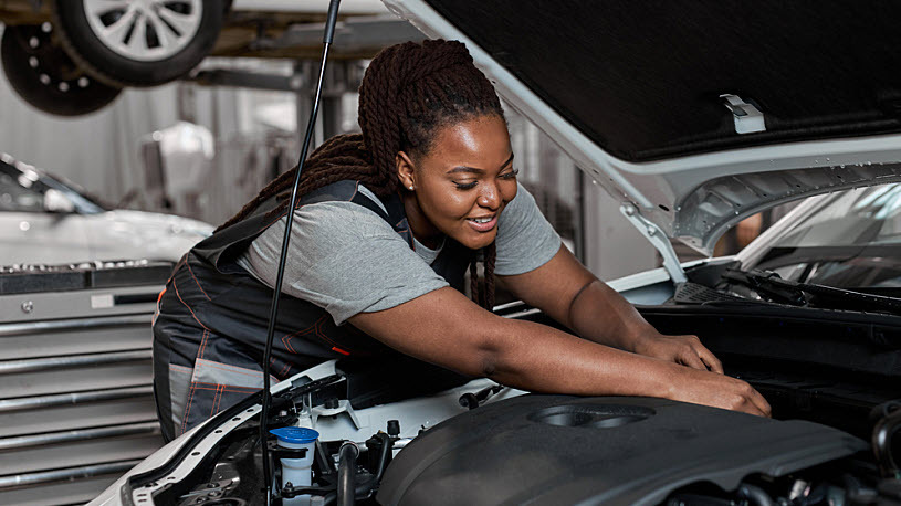 Young female car repair worker checks oil level in car engine Young Black female car repair worker checks the oil level in car engine, side view